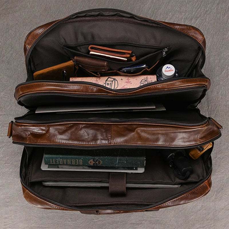 An open Leather 17" Business Travel Briefcase reveals neatly organized contents, including a wallet, sunglasses, a laptop, a pen, a book titled "BERNARDT," a small container, and other personal items. This business travel bag features multiple compartments and appears well-constructed.