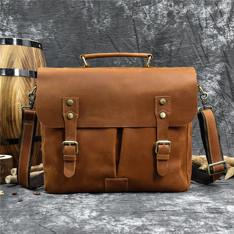 The Leather 14" Briefcase Shoulder Bag, featuring a vintage-style brown leather with brass buckles and a top handle, is displayed against a textured gray background. A wooden barrel is partially visible on the left side of the image. This genuine cowhide bag includes an adjustable shoulder strap and showcases a professional, classic design.