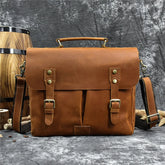 A brown Leather 14" Briefcase Shoulder Bag with brass buckle closures and a top handle, crafted from genuine cowhide. It features an adjustable shoulder strap and is set against a textured gray background. To the left, there is a wooden barrel, adding a rustic element to the professional style scene.