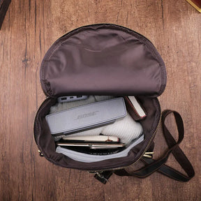 The Vintage Leather Travel Backpack, crafted from genuine cowhide leather, sits on a wooden surface with a portable speaker, smartphone, and folded clothing inside—ideal for the stylish traveler.