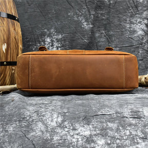 The image shows the bottom view of a Leather 14" Briefcase Shoulder Bag made from genuine cowhide brown leather, placed on a textured gray surface. The briefcase features visible stitching along the edges. To the left, parts of a wooden barrel and a rope are partially visible. The backdrop consists of distressed fabric, adding a touch of rustic charm.