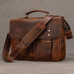 The Leather 14" Briefcase Shoulder Bag, made of genuine cowhide in a rich brown color, is displayed against a plain background. This satchel features a top handle and an adjustable shoulder strap. It boasts two front buckle closures, side pockets, and stitching accents. The leather has a distressed, vintage look that blends professional style with timeless charm.