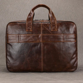 The Leather 17" Business Travel Briefcase offers a genuine leather construction with two sturdy handles and a zippered top. Its sleek design boasts visible stitching, a horizontal band detail on the front, and specialized laptop compartments. Ideal for business travel, it is showcased against a plain gray background.