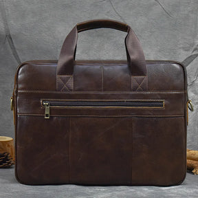 A 16" Brown Leather Briefcase, crafted from genuine leather, features dual handles and a zippered front pocket, elegantly displayed against a gray fabric background. This briefcase, which also serves as a sophisticated handbag, showcases a sleek minimalist design with stitched detailing and metal hardware on the sides. An adjustable strap adds to its convenience.