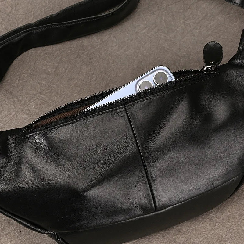 A Leather Belt Bag rests on a textured surface. The main compartment is partially unzipped, revealing a modern smartphone with dual rear camera lenses inside. Crafted from genuine cowhide leather, the strap of the fanny pack is partially visible, showcasing durable alloy hardware.