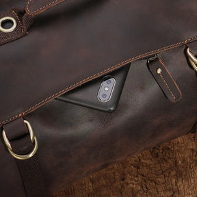 The Vintage Leather Travel Backpack, crafted from genuine cowhide leather and accented with metal rings and buckles, is slightly open to reveal a smartphone with dual cameras inside. This stylish traveler's essential rests elegantly on a wooden surface.