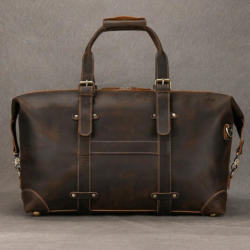 The Weekender 20 Leather Travel Duffle is a large, dark brown leather bag featuring sturdy straps and metal buckles. Crafted from genuine cowhide leather, it comes with a top handle and includes a removable strap. The minimalist design with visible stitching stands out against the plain gray background.