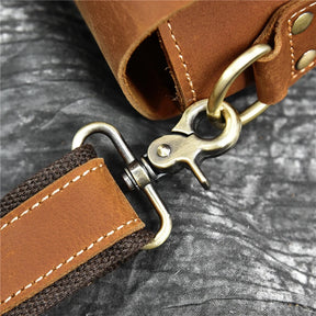 Close-up shot of the Leather 14" Briefcase Shoulder Bag strap attached to a metal ring with a clasp. The stitching on the genuine cowhide is visible, and the strap includes a combination of leather and dark brown fabric. The background is a textured dark surface, enhancing its professional style.