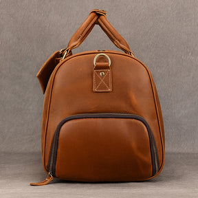 The Weekender Duffle 22 Leather Travel Bag is a small, brown leather backpack crafted from genuine cowhide leather. It features a front zippered pocket, a top handle, and an adjustable shoulder strap. Additionally, the bag includes a metal ring attached to a leather patch near the top. The background consists of a plain grey surface.