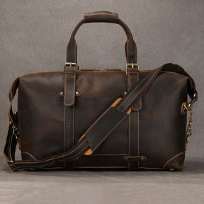 The Weekender 20 Leather Travel Duffle is showcased against a gray background, highlighting its large size and dark brown genuine cowhide leather construction. This travel bag features sturdy handles, a removable shoulder strap, white stitching, and brass hardware that lend it a vintage, rugged appearance.