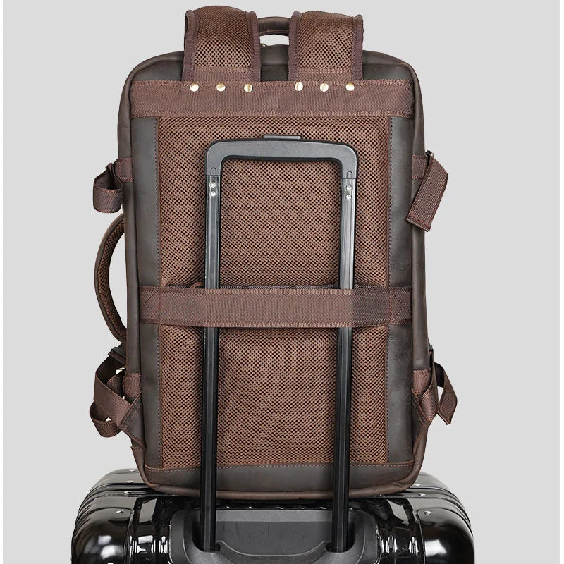 The Vintage Leather Travel Backpack, featuring a vintage style and crafted from genuine leather with a fabric texture, elegantly rests secured to the handle of a black suitcase. This brown backpack includes shoulder straps, a luggage strap, and multiple pockets, all set against a neutral gray background.