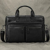 The 15" Black Briefcase is crafted from genuine leather and includes two top handles along with an adjustable strap. The front boasts multiple zippered compartments, featuring two smaller zip pockets and one larger zipper pocket above. Ideal as a professional handbag, it complements a textured, muted gray surface beautifully.