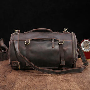 A Vintage Leather Travel Backpack made from genuine cowhide leather, complete with brass buckles and adjustable straps, rests on a wooden surface. Ideal for the stylish traveler, it is accompanied by the backdrop of a red analog clock and part of a decorative object.