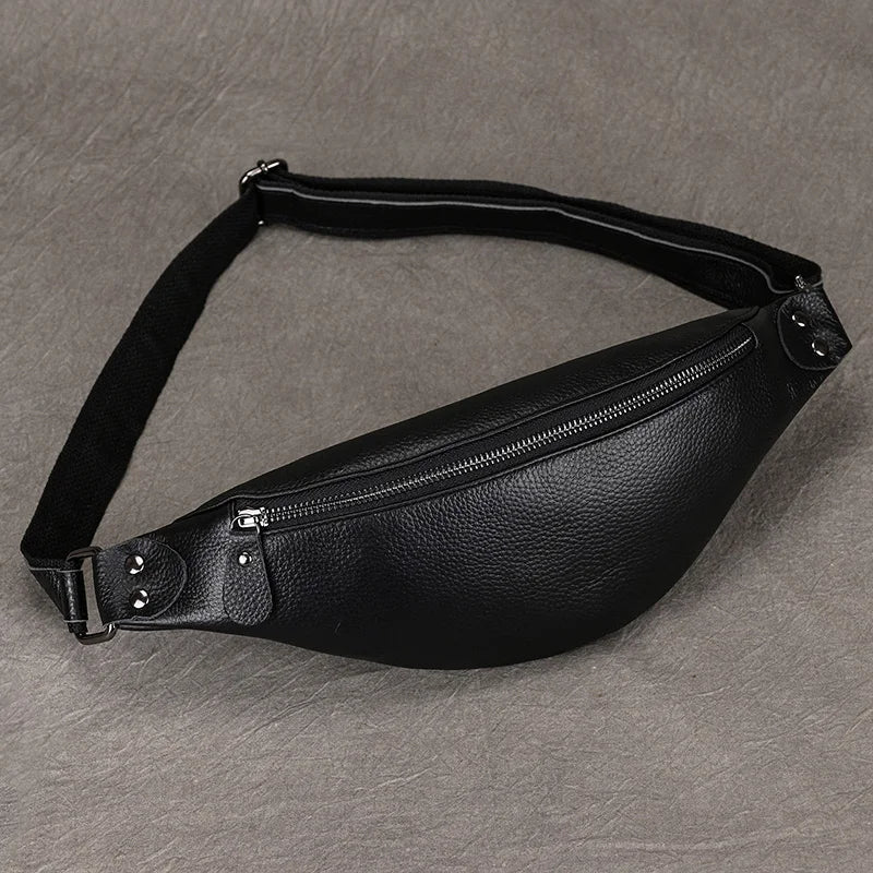 A Black Leather Belt Bag with a zippered main compartment and an adjustable strap lies on a textured gray surface. The fanny pack, crafted from premium leather, features metal studs near the zipper and a cloth strap buckle for fastening.