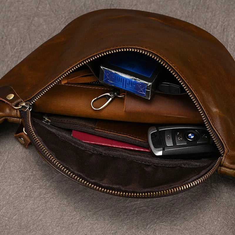 A Leather Belt Bag sits open, revealing its contents. Inside, there is a key fob, a set of keys with durable alloy hardware, a pack of cigarettes, a brown leather wallet, and a pen. The bag is placed on a gray textured surface.