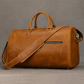 The Leather Travel Duffle is a large, brown genuine leather bag with vintage charm, showcased against a gray background. It includes two handles, a detachable shoulder strap, a front zipper pocket, and metal feet for durability.