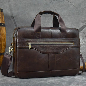 The 16" Brown Leather Briefcase is showcased against a gray, textured background. This genuine leather handbag, with its rich and polished finish, features sturdy handles, a front zippered pocket, and an adjustable shoulder strap attached by gold-colored clasps.