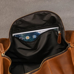 The open Weekender Duffle 22 Leather Travel Bag, crafted from genuine brown cowhide leather, reveals a zippered internal pocket holding a tablet and other items. The tablet screen shows various app icons against the bag's stylish black fabric interior.