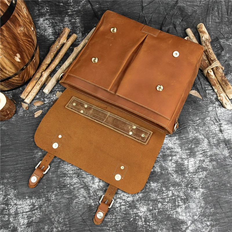 The Leather 14" Briefcase Shoulder Bag, crafted from genuine brown cowhide, features an open front flap that reveals multiple compartments and pockets. Displayed on a textured gray surface with wooden sticks and a wooden bucket in the background, this professional-style bag is highlighted by its buckle straps and gold-colored hardware.