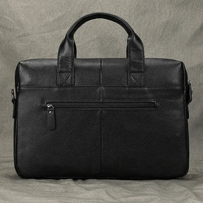 The 15" Black Briefcase rests against a textured gray background, featuring dual handles, an adjustable strap, and a front zippered pocket. Made from genuine leather, it showcases a smooth, polished appearance with detailed stitching.