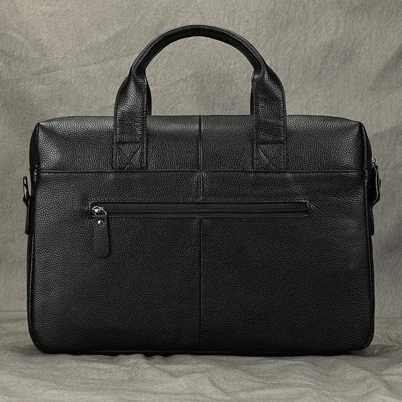 The 15" Leather Briefcase in black genuine leather offers a slim, lightweight design with sturdy handles and a zippered front pocket, complemented by a textured finish. Resting on a gray surface, this professional handbag exudes a sleek and polished appearance.