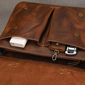 A close-up of an open Leather 14" Briefcase Shoulder Bag reveals its professional style. Inside the genuine cowhide bag, there are multiple compartments holding various items including a white power bank, a keychain with keys attached, and a mobile phone. The bag has metal snap buttons for closure.