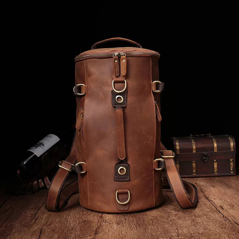 The Vintage Leather Travel Backpack, made from authentic cowhide leather featuring buckles and straps, is showcased on a wooden surface. A wine bottle and a small chest in the background contribute to a rustic ambiance ideal for the fashionable traveler.