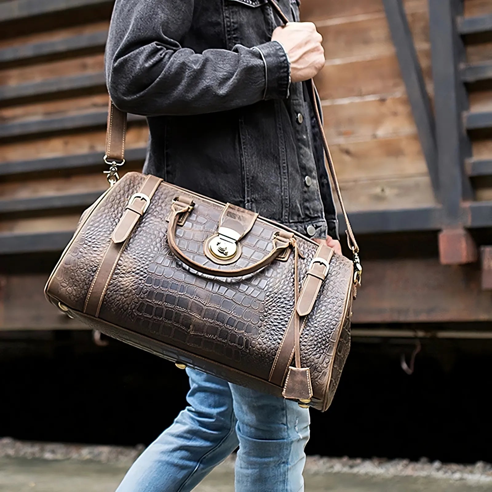 A person wearing a dark jacket and blue jeans carries the Crocodile Pattern Leather Travel Bag, featuring a brown genuine leather with a crocodile texture and gold accents. The bag, boasting a spacious interior, includes both a shoulder strap and handles. In the background stands a wooden structure, possibly part of a train or building.